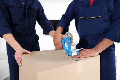 Photo of Male movers packing box with adhesive tape indoors, closeup