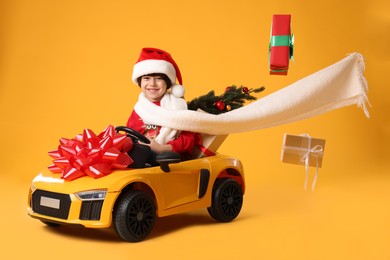 Photo of Cute little boy in Santa hat with Christmas tree and gift boxes driving children's electric toy car on yellow background