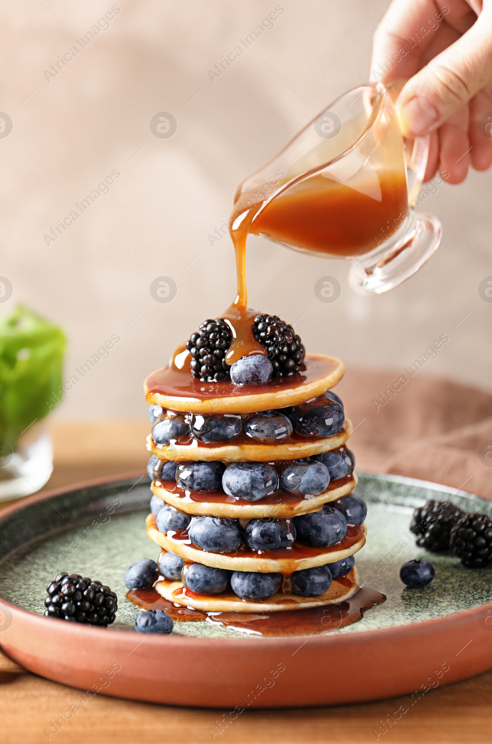 Photo of Woman pouring syrup onto tasty pancakes with berries on plate, closeup