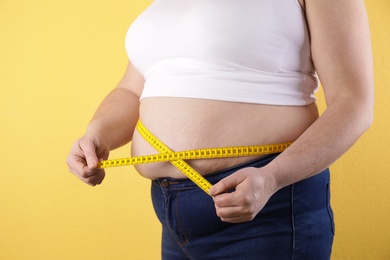 Overweight woman measuring waist with tape on color background, closeup