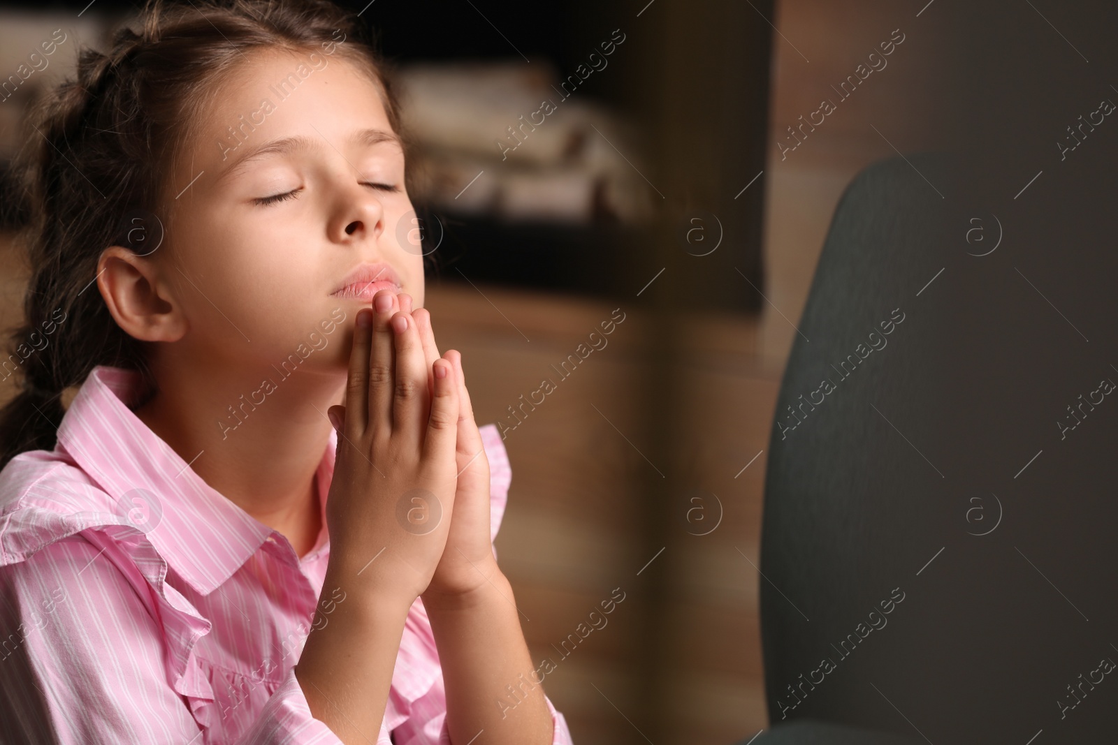 Photo of Cute little girl with hands clasped together praying at home. Space for text