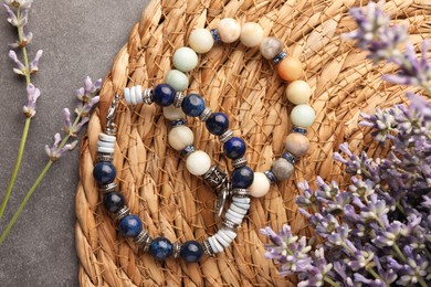 Photo of Beautiful bracelets with gemstones, lavender and wicker mat on grey table, flat lay