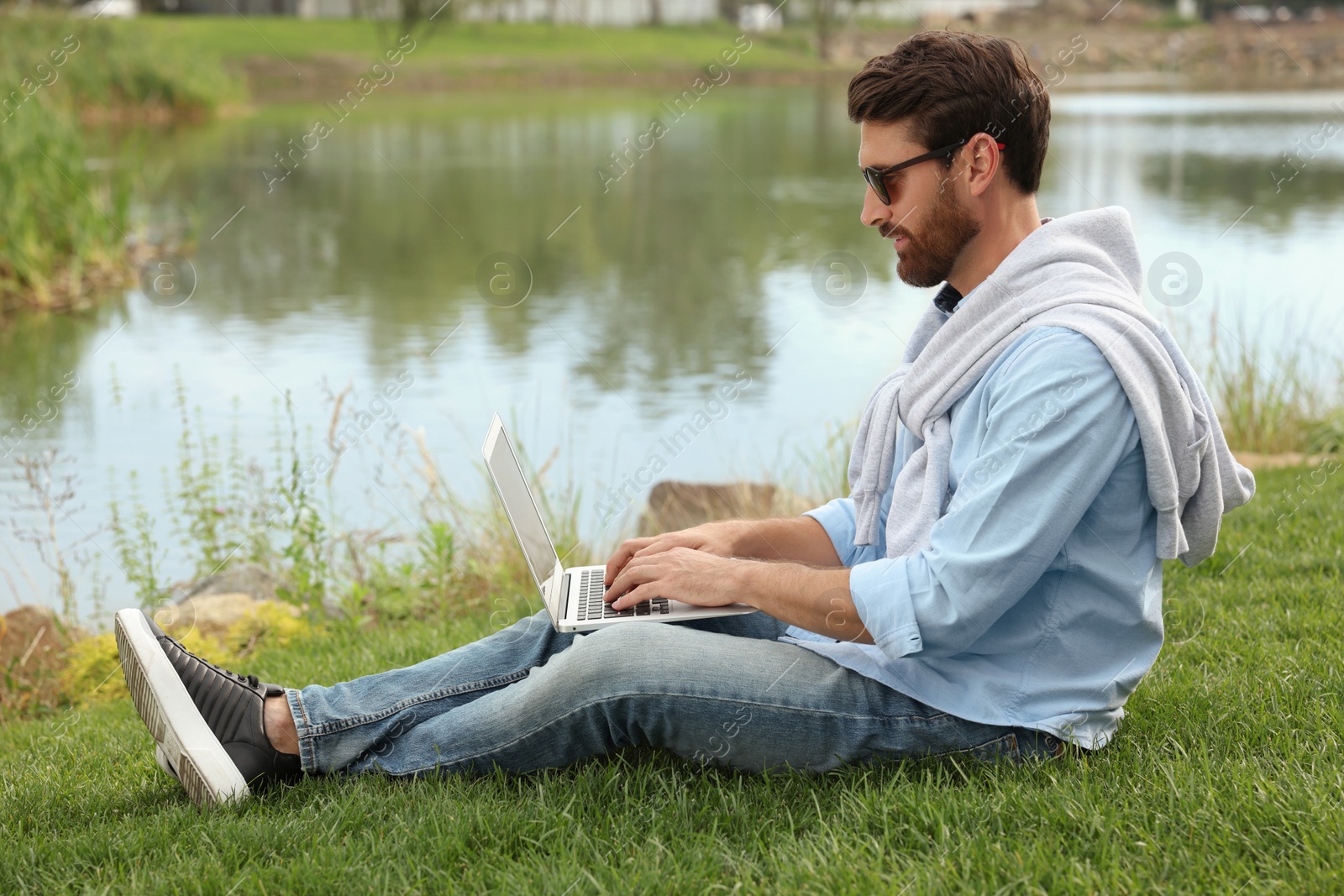 Photo of Man in sunglasses with laptop on green grass near lake