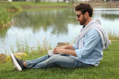 Man in sunglasses with laptop on green grass near lake
