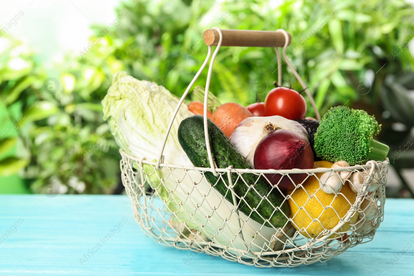 Photo of Fresh vegetables in metal basket on light blue wooden table against blurred green background