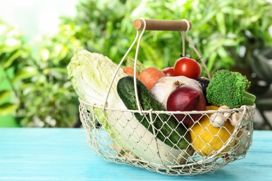 Photo of Fresh vegetables in metal basket on light blue wooden table against blurred green background
