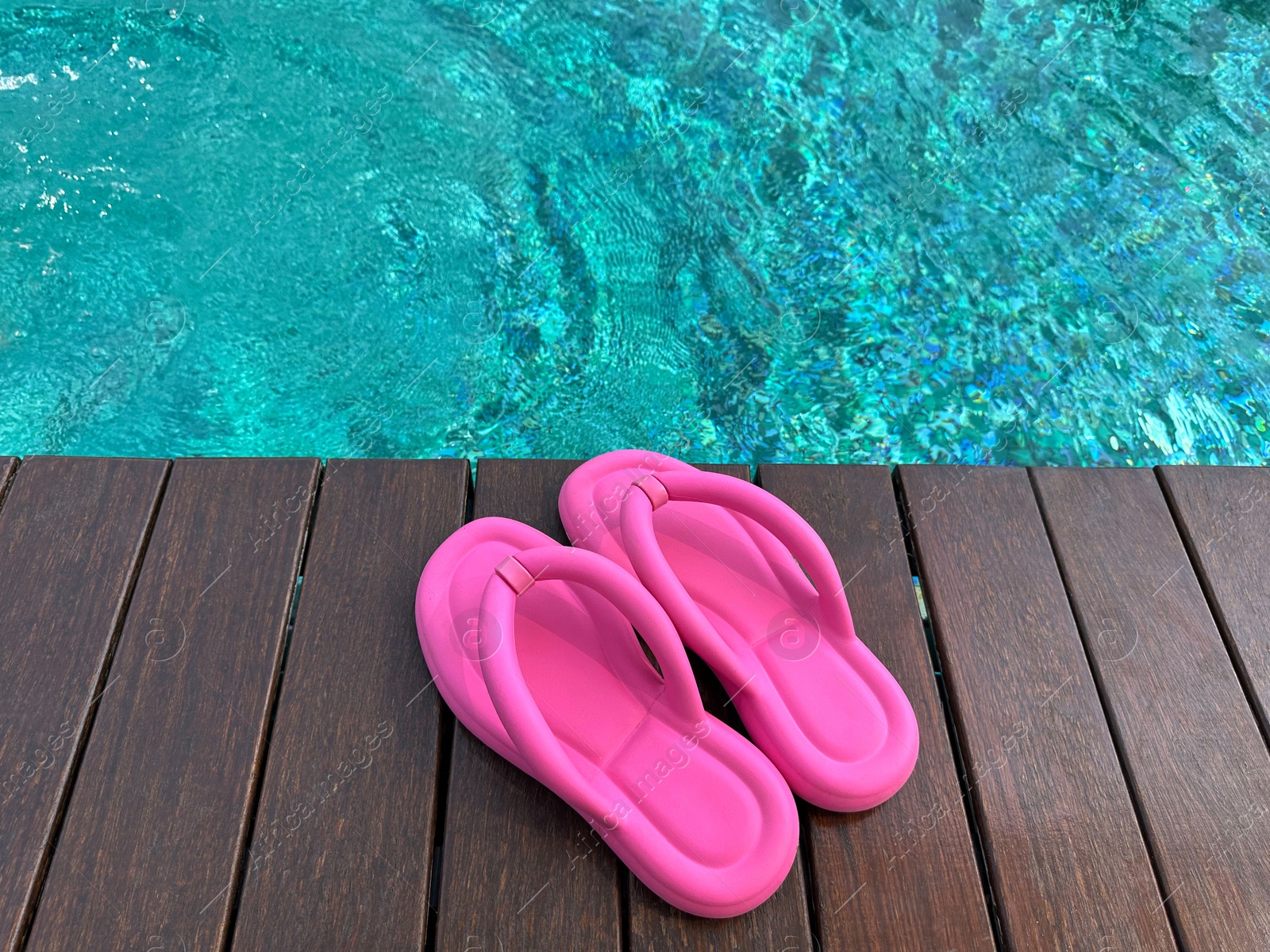 Photo of Clear rippled water in swimming pool and pink flip-flops on wooden deck outdoors, closeup