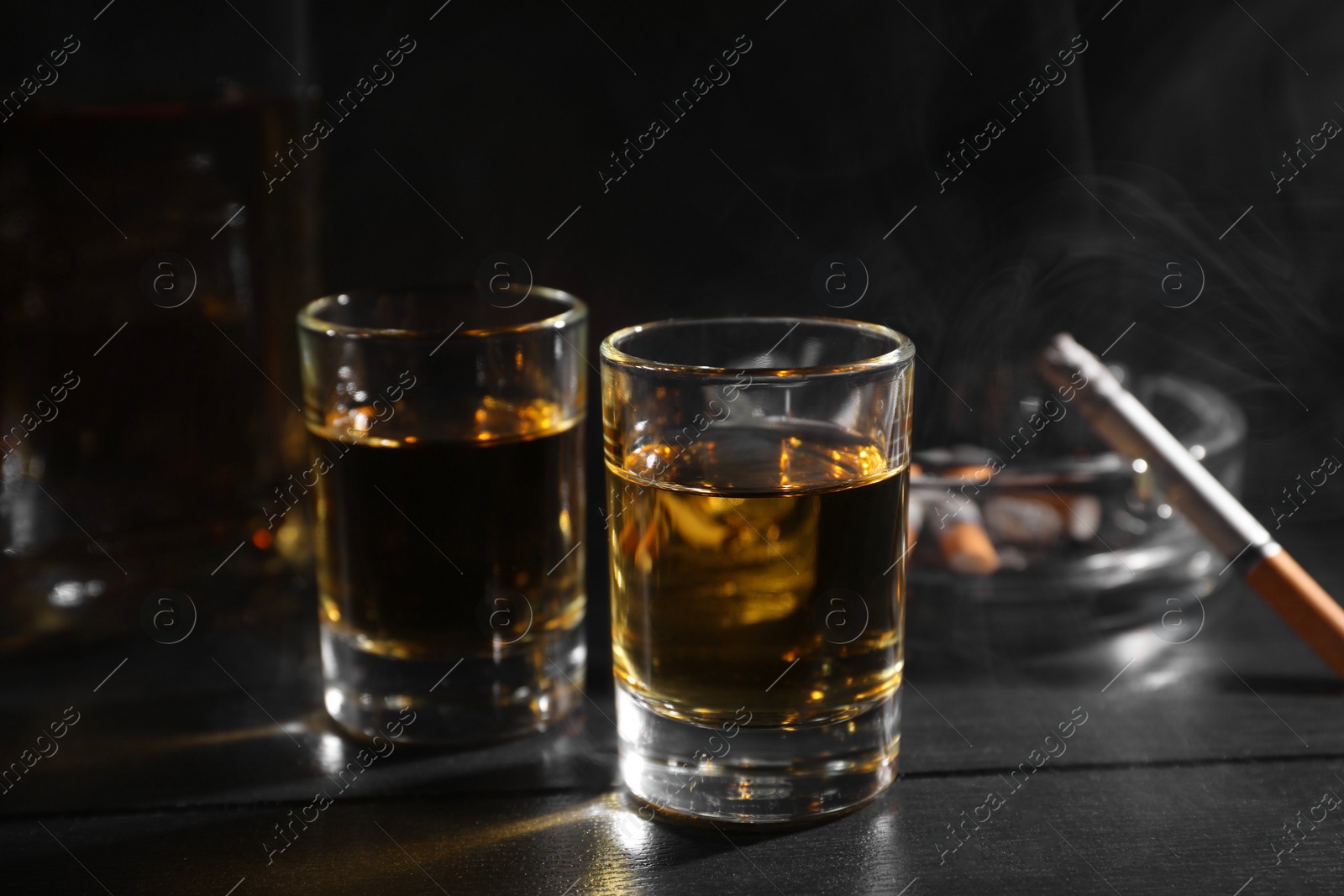 Photo of Alcohol addiction. Whiskey in glasses, smoldering cigarette and ashtray on black wooden table