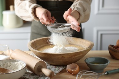 Photo of Making dough. Woman sifting flour into bowl at wooden table in kitchen, closeup