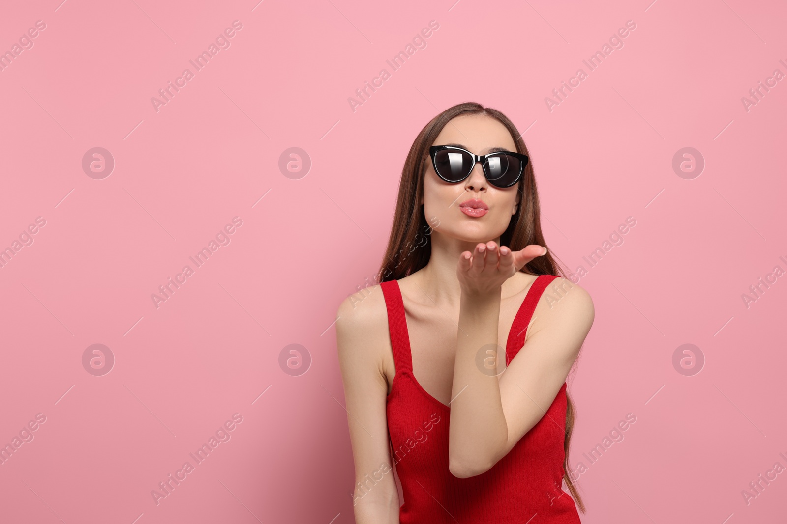 Photo of Beautiful young woman with sunglasses blowing kiss on pink background, space for text