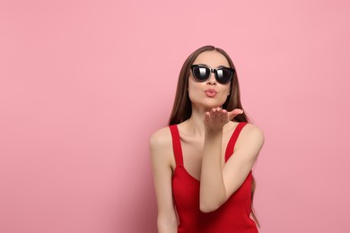 Photo of Beautiful young woman with sunglasses blowing kiss on pink background, space for text
