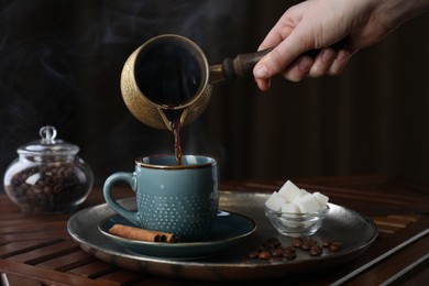 Turkish coffee. Woman pouring brewed beverage from cezve into cup at wooden table, closeup