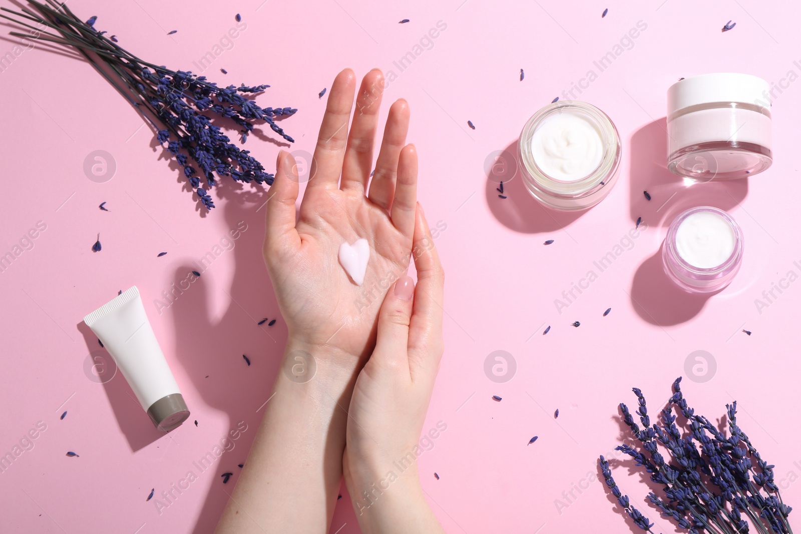 Photo of Woman applying hand cream and lavender flowers on pink background, top view