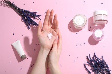 Woman applying hand cream and lavender flowers on pink background, top view