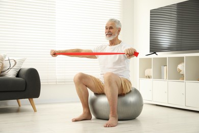 Photo of Senior man doing exercise with elastic resistance band on fitness ball at home