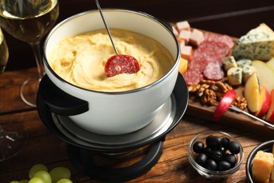 Photo of Fork with piece of sausage, melted cheese in fondue pot and other products on wooden table, closeup