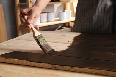 Photo of Man with brush applying wood stain onto wooden surface indoors, closeup