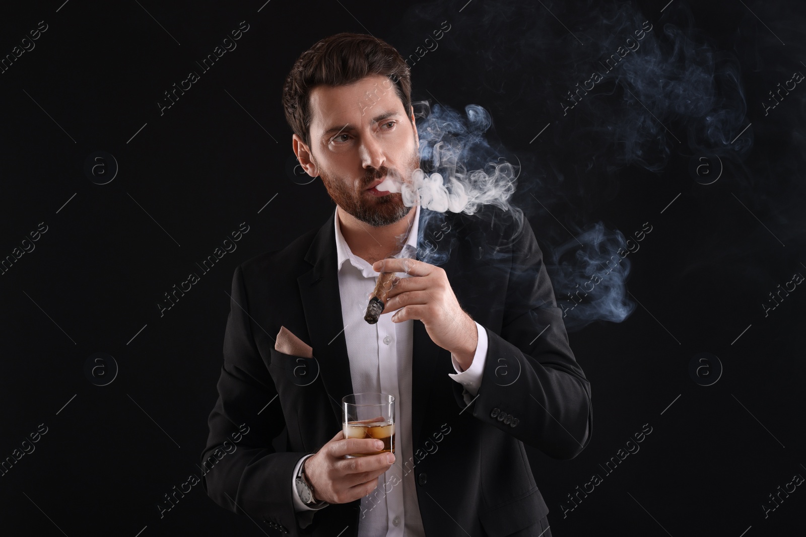 Photo of Handsome man in elegant suit with glass of whiskey smoking cigar on black background