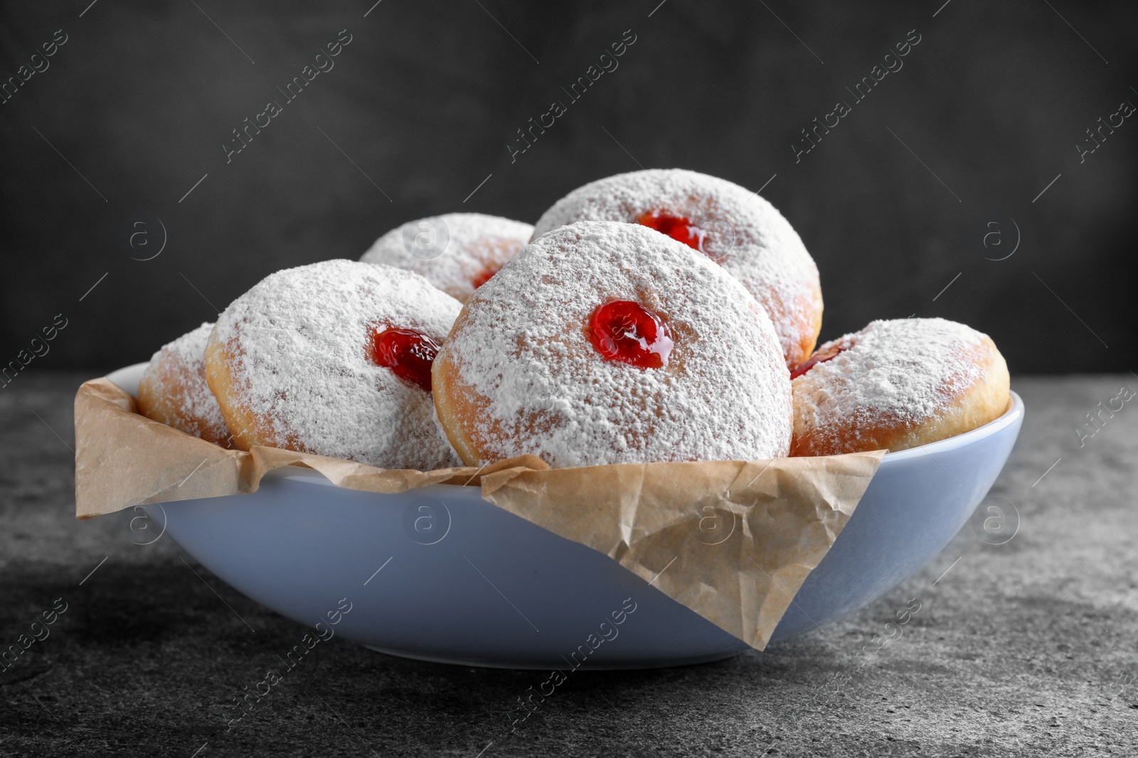 Photo of Delicious donuts with jelly and powdered sugar in bowl on grey table