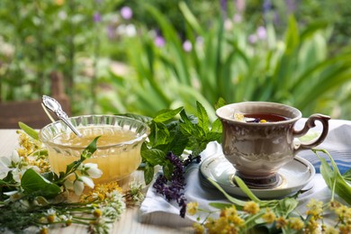 Photo of Cup of hot aromatic tea, honey and different fresh herbs on white wooden table outdoors