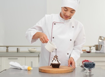 Photo of Female pastry chef pouring chocolate sauce onto cupcake at table in kitchen