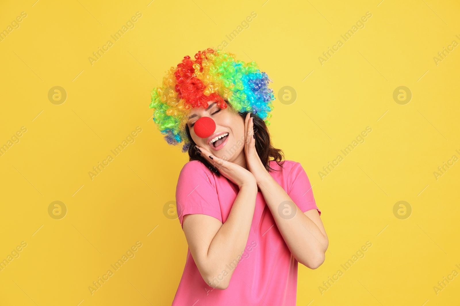 Photo of Joyful woman with rainbow wig and clown nose on yellow background. April fool's day