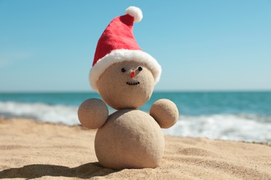 Photo of Snowman made of sand with Santa hat on beach near sea, closeup. Christmas vacation