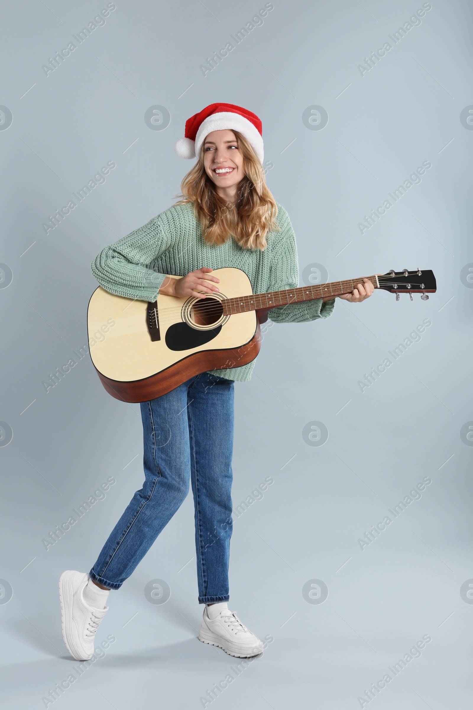 Photo of Young woman in Santa hat playing acoustic guitar on light grey background. Christmas music