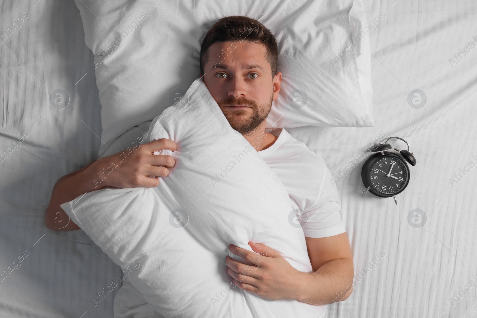 Photo of Sleepy man with pillow and alarm clock in bed, top view. Insomnia problem