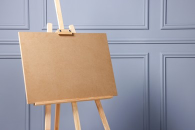 Wooden easel with blank board near grey wall indoors, closeup. Space for text