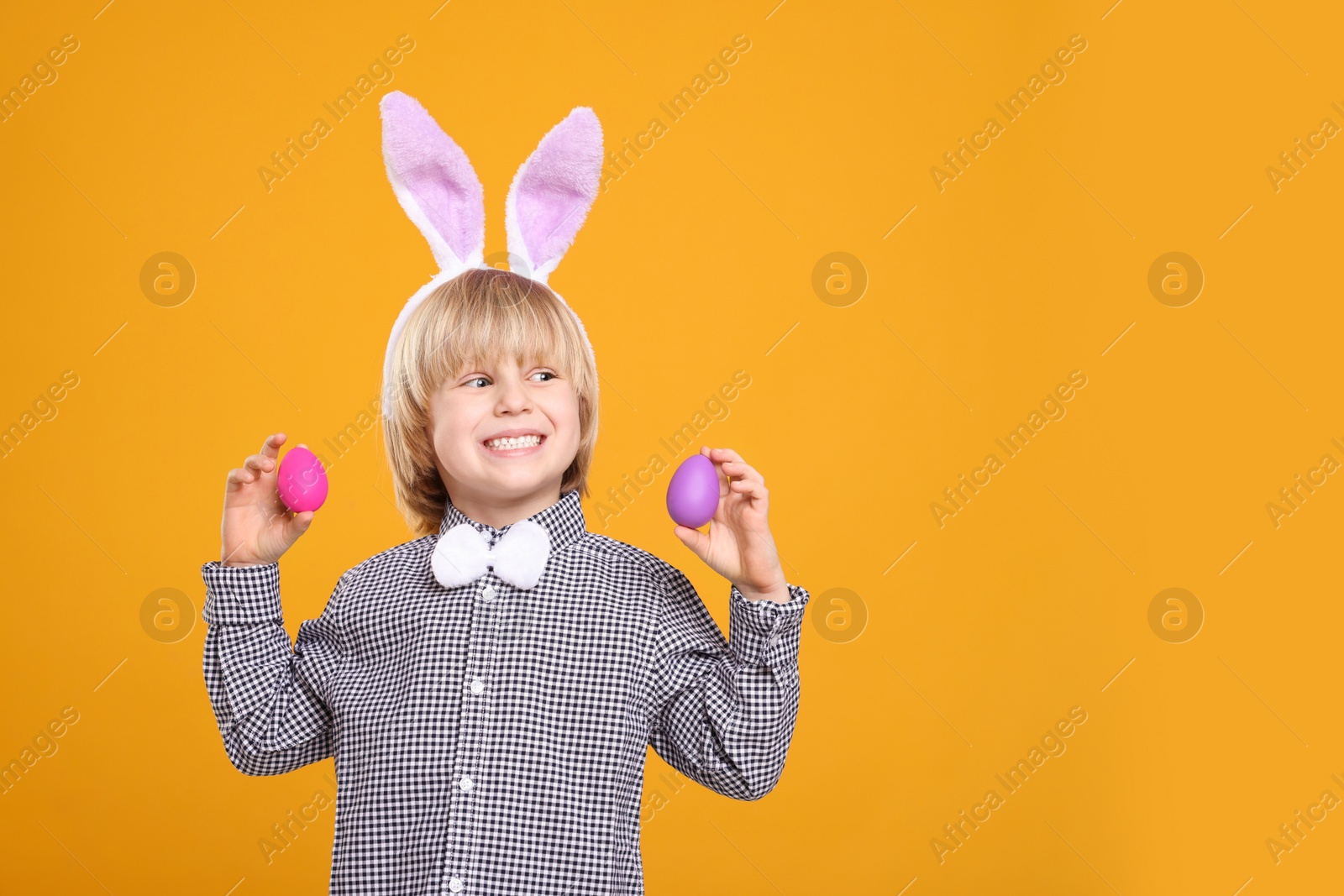 Photo of Happy boy in bunny ears headband holding painted Easter eggs on orange background. Space for text