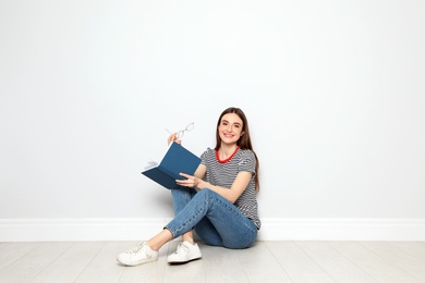 Young woman reading book on floor near white wall, space for text