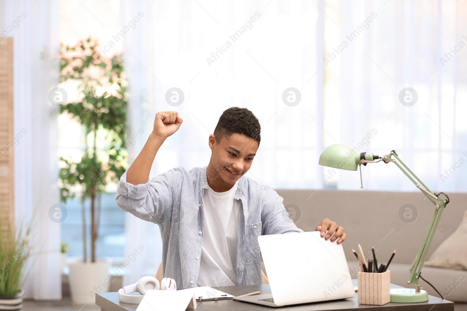 Photo of Emotional African-American teenage boy with laptop at table in room