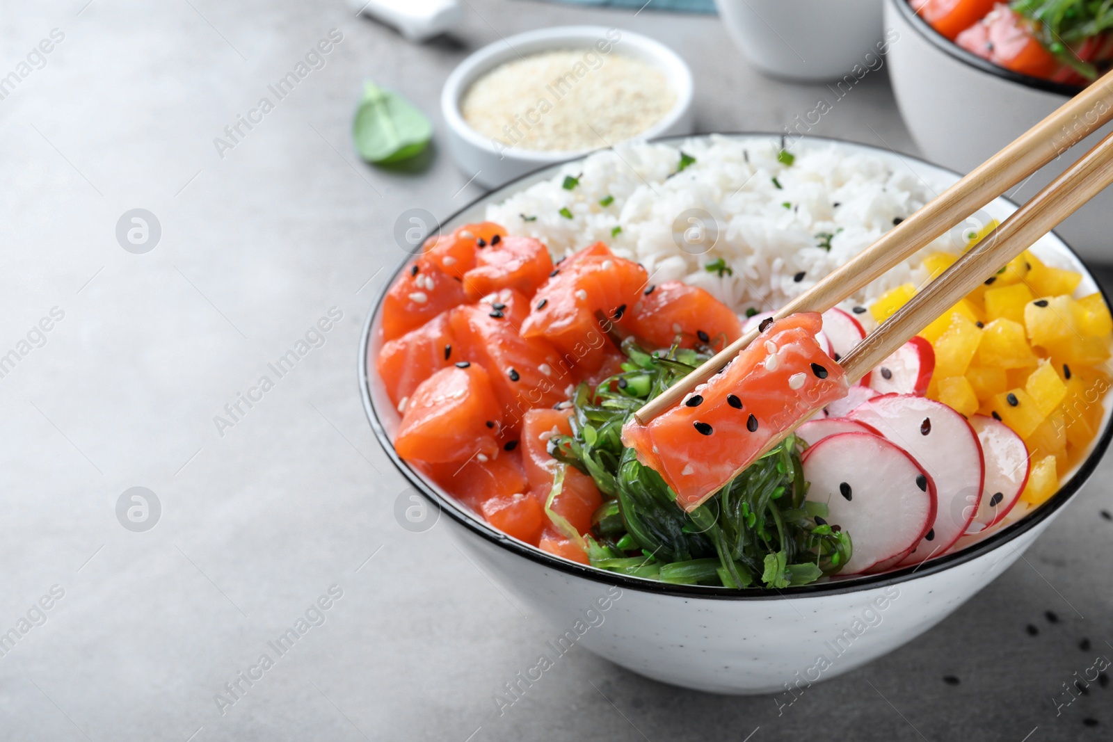 Photo of Wooden chopsticks with piece of salmon over delicious poke bowl on light grey table, closeup. Space for text