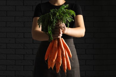 Woman holding fresh ripe juicy carrots against dark brick wall, closeup
