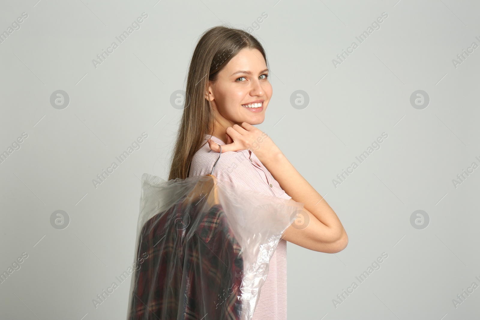 Photo of Young woman holding hanger with shirt in plastic bag on light grey background. Dry-cleaning service