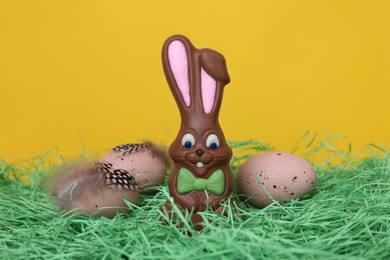 Photo of Easter celebration. Cute chocolate bunny and painted eggs with feathers on grass against yellow background