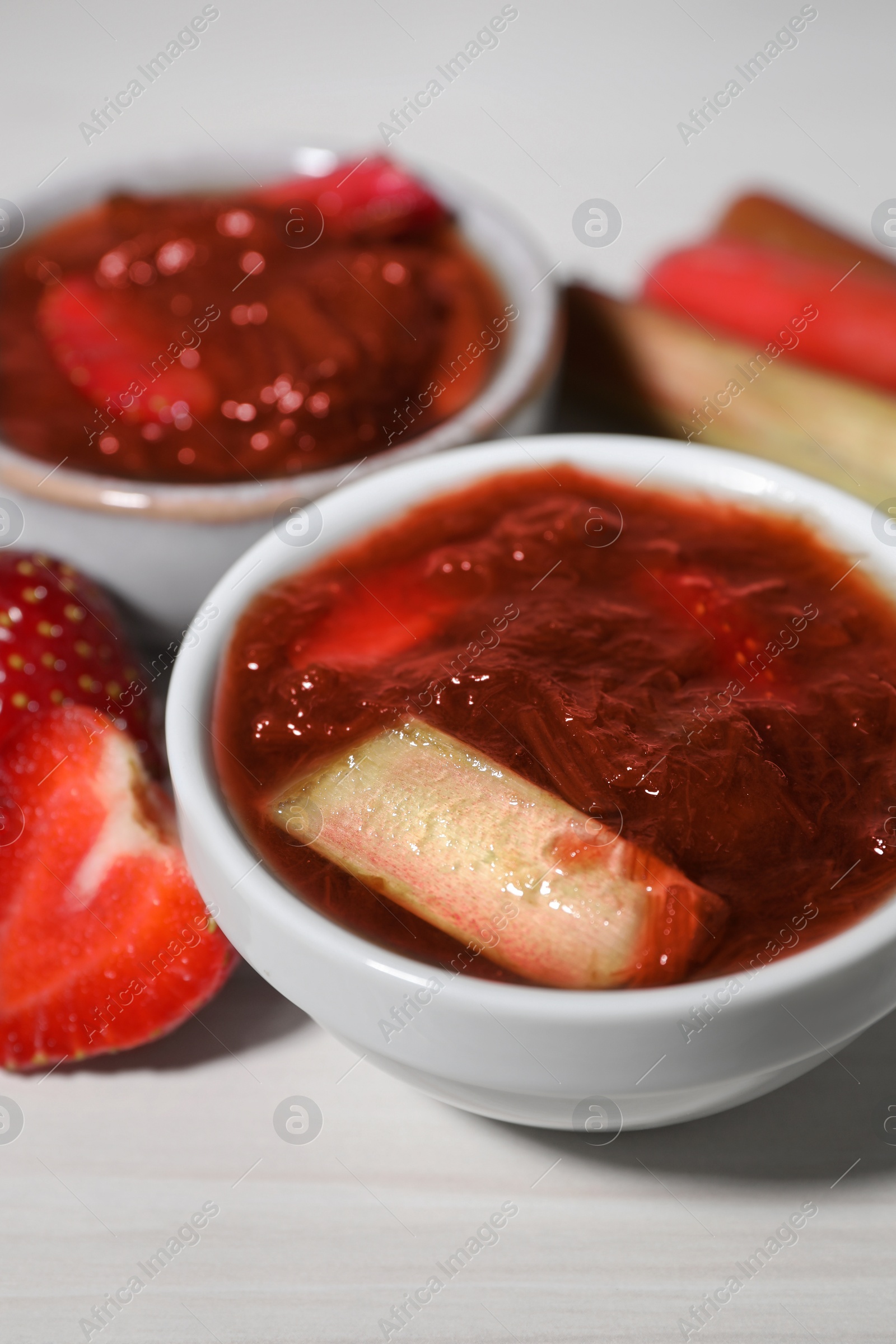 Photo of Tasty rhubarb jam in bowls and strawberries on white wooden table, closeup