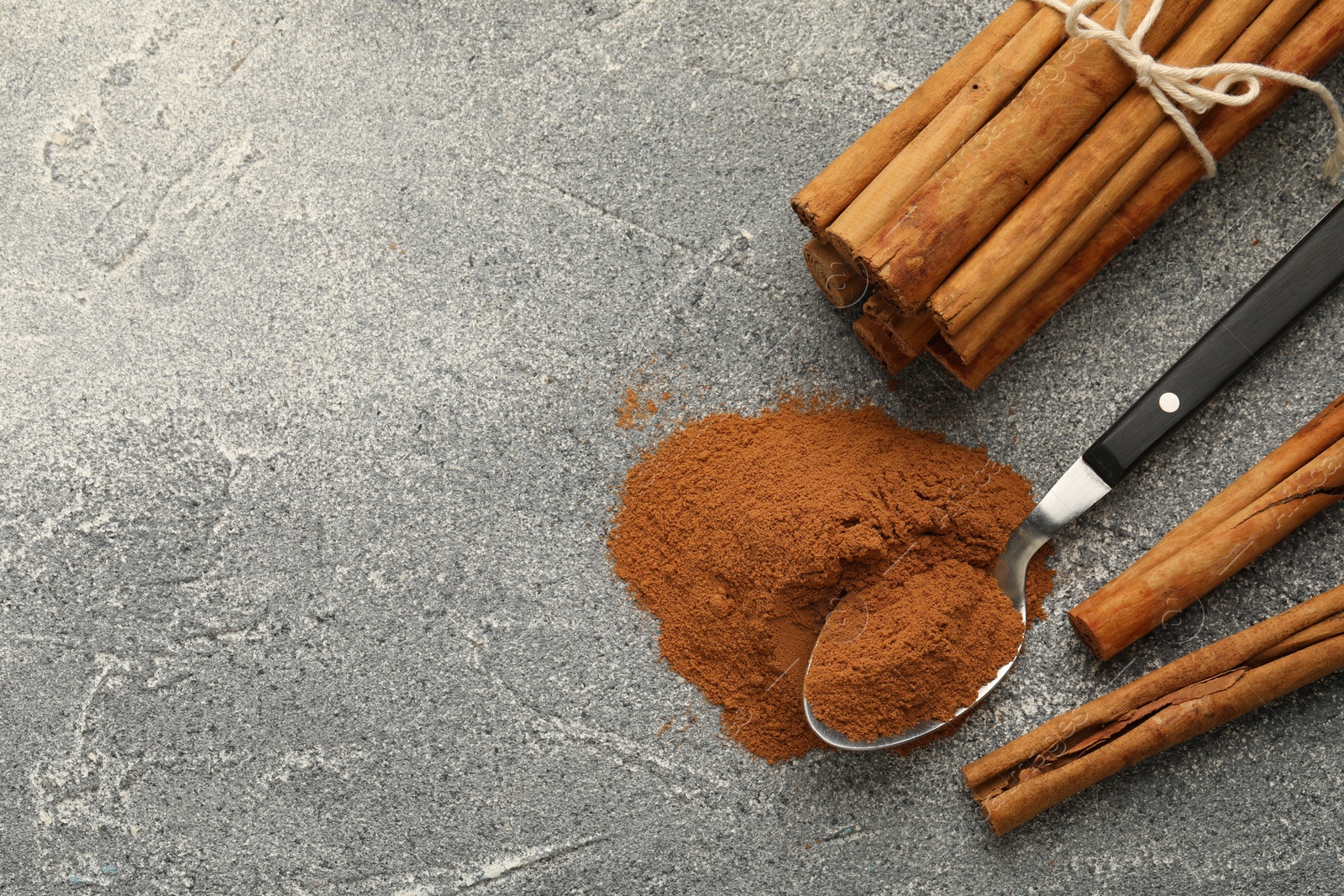 Photo of Spoon with cinnamon powder and sticks on grey table, flat lay. Space for text