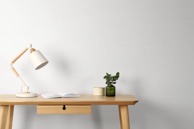 Photo of Stylish modern desk lamp, open book and plant on wooden table near white wall, space for text
