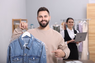 Photo of Dry-cleaning service. Happy man holding hanger with denim jacket in plastic bag indoors. Worker using laptop at workplace