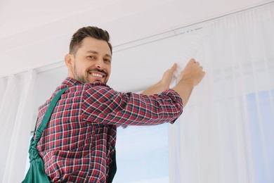 Photo of Worker in uniform hanging window curtain indoors, low angle view