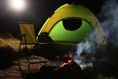 Photo of Smoking bonfire and folding chairs near camping tent outdoors at night