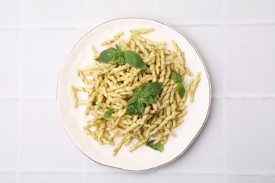 Plate of delicious trofie pasta with pesto sauce and basil leaves on white tiled table, top view