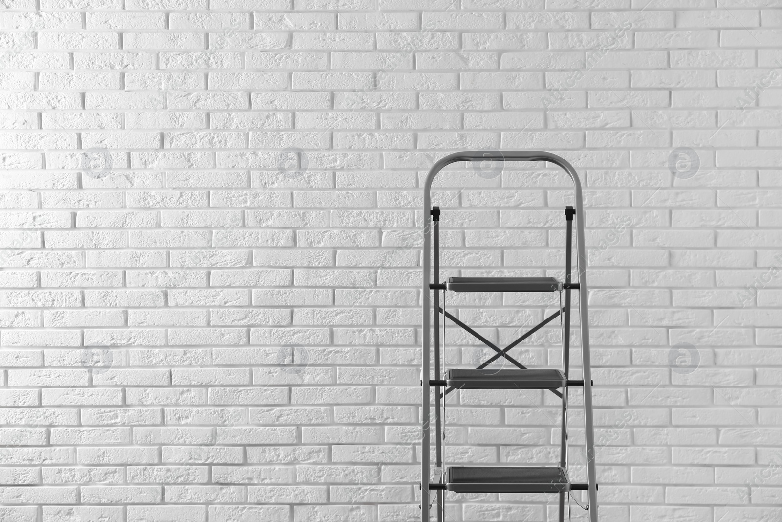 Photo of Modern metal stepladder against white brick background. Space for text