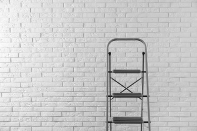 Photo of Modern metal stepladder against white brick background. Space for text