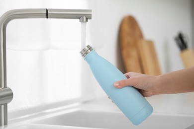 Woman pouring fresh water from tap into thermo bottle in kitchen, closeup