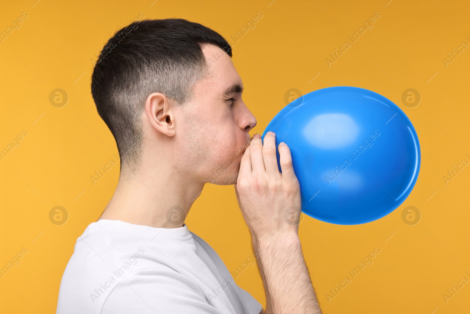 Photo of Young man inflating light blue balloon on yellow background