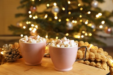 Cups of tasty cocoa with marshmallows and pine cone on wooden tray near Christmas tree
