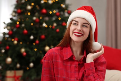 Beautiful young woman in Santa hat near Christmas tree at home, space for text